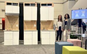 Two women stand next to modular kitchen cabinets. 