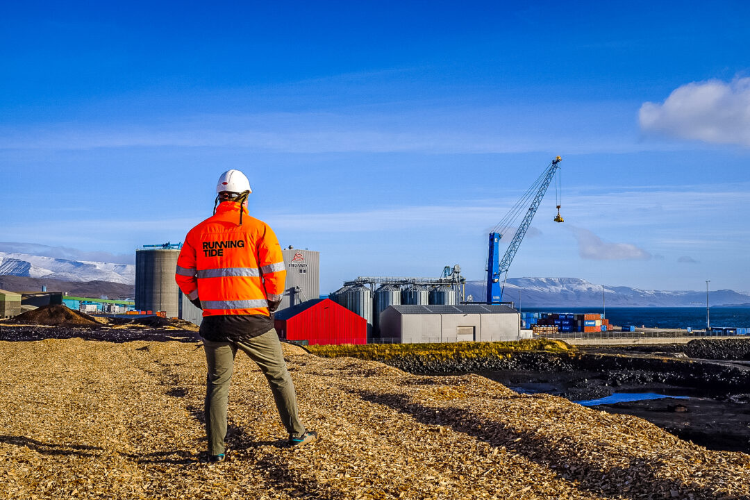 A man in an orange jacket and hardhat stands on the coast by a crane.