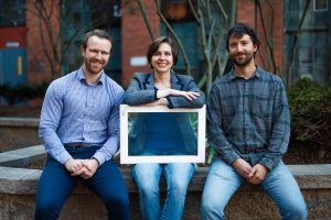 Three people sit on an outside bench and smile at the camera while holding a framed piece of glass.