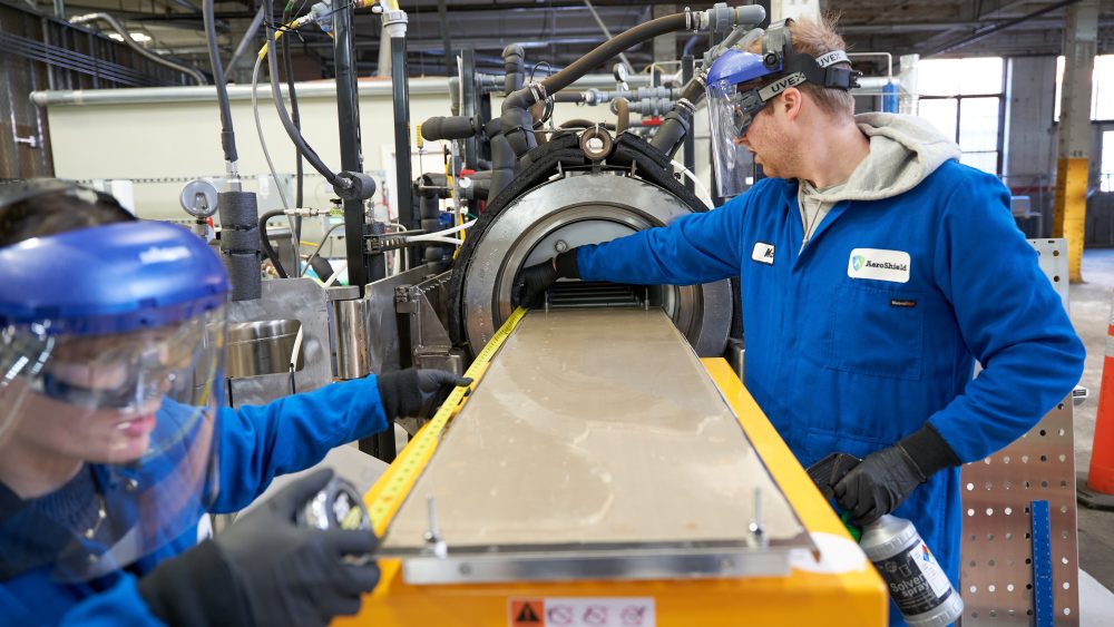 Two people in blue lab coats and face shields work on a piece of equipment producing a sheet of glass.