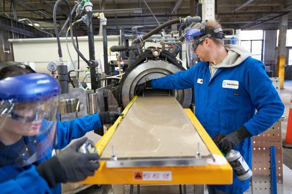 Two people in blue lab coats and face shields work on a piece of equipment producing a sheet of glass.