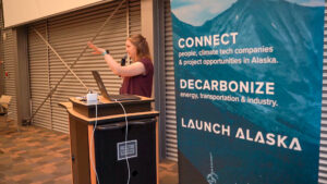 A woman speaks into a microphone at a podium next to a sign reading Connect, Decarbonize, Launch Alaska.