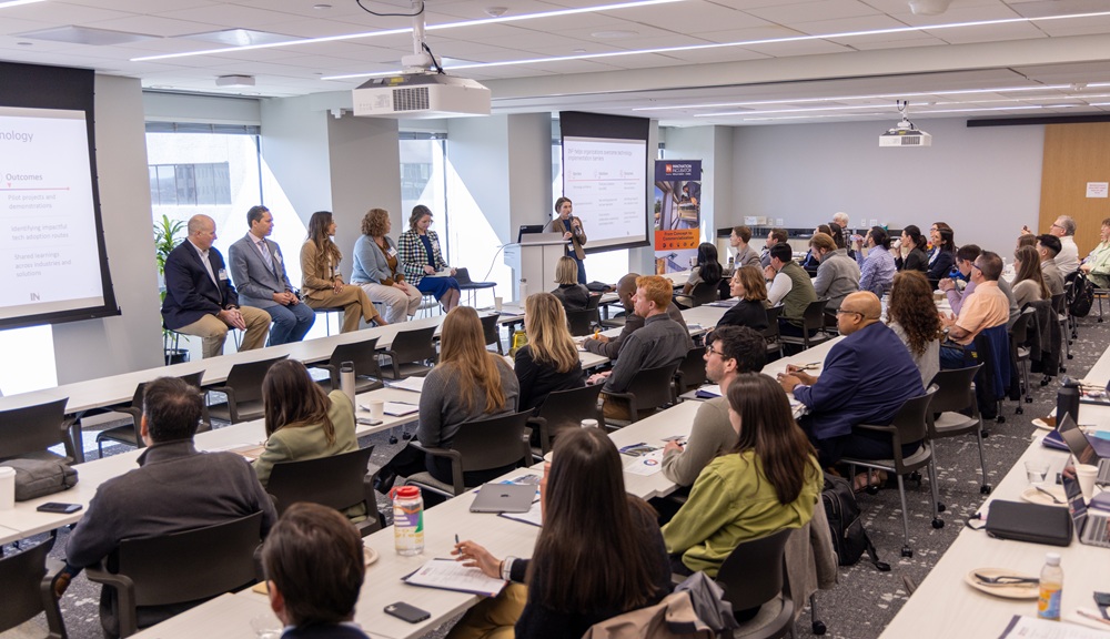 A meeting room full of people listens to a panel of five people speak.