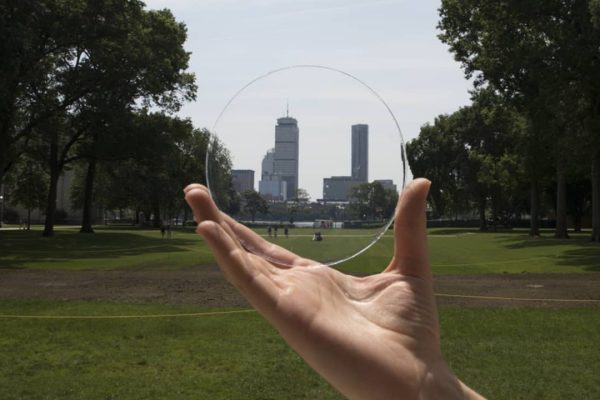 A hand holds a clear, round glass disc in a park, with a city skyline visible in the background through the transparent glass.