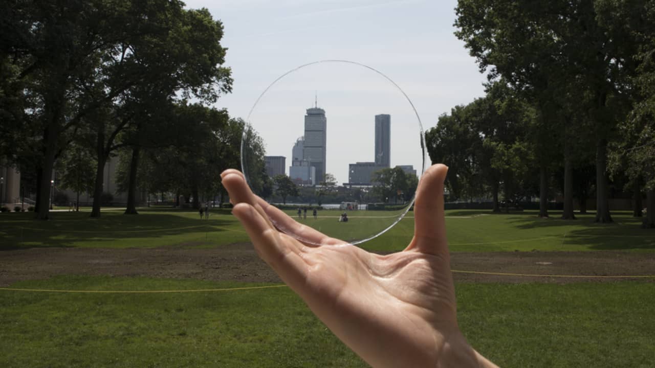 A hand holds a clear, round glass disc in a park, with a city skyline visible in the background through the transparent glass.
