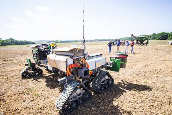 A robotic machine sits in a dirt field while a group of four people stands in the background looking.