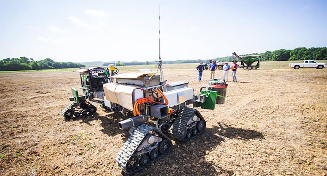A robotic machine sits in a dirt field while a group of four people stands in the background looking.