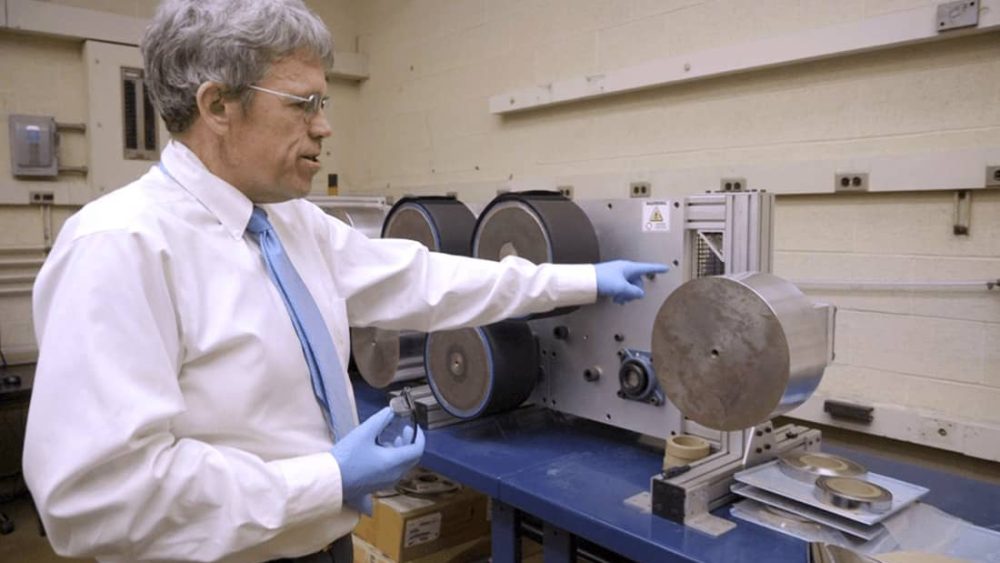 A man wearing gloves gestures at a machine with multiple rollers in a workshop setting.