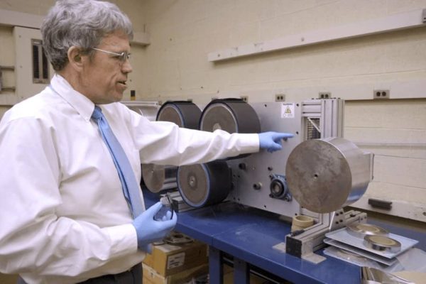 A man wearing gloves gestures at a machine with multiple rollers in a workshop setting.