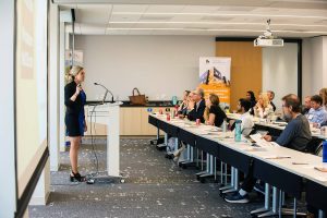 A woman presents slides to a meeting room of people.