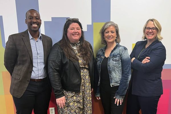 Four people stand in front of a wall, posing and smiling for the camera.