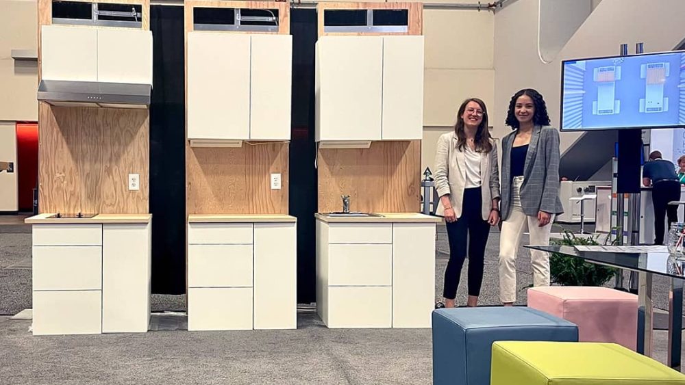 Two women stand next to modular kitchen cabinets.