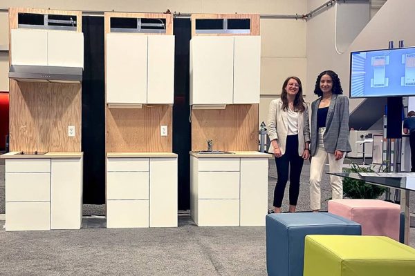 Two women stand next to modular kitchen cabinets.