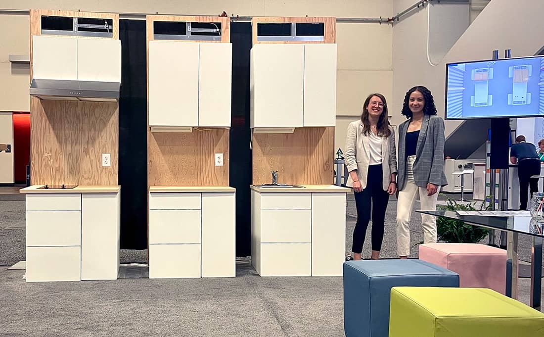 Two women stand next to modular kitchen cabinets.
