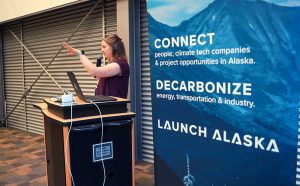 A woman speaks into a microphone at a podium next to a sign reading Connect, Decarbonize, Launch Alaska.