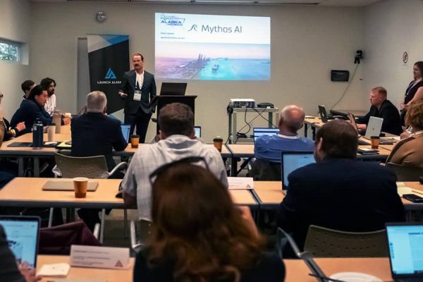 People sit at tables listening to a man in a suit give a presentation.