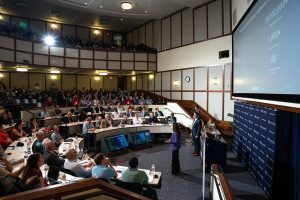 A large auditorium meeting room full of people watching two presenters.