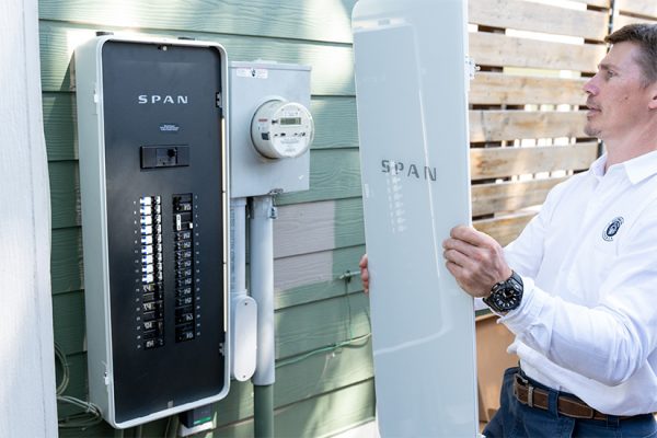 A man holds the cover of a power panel next to the panel which is hanging on the outside of a home.