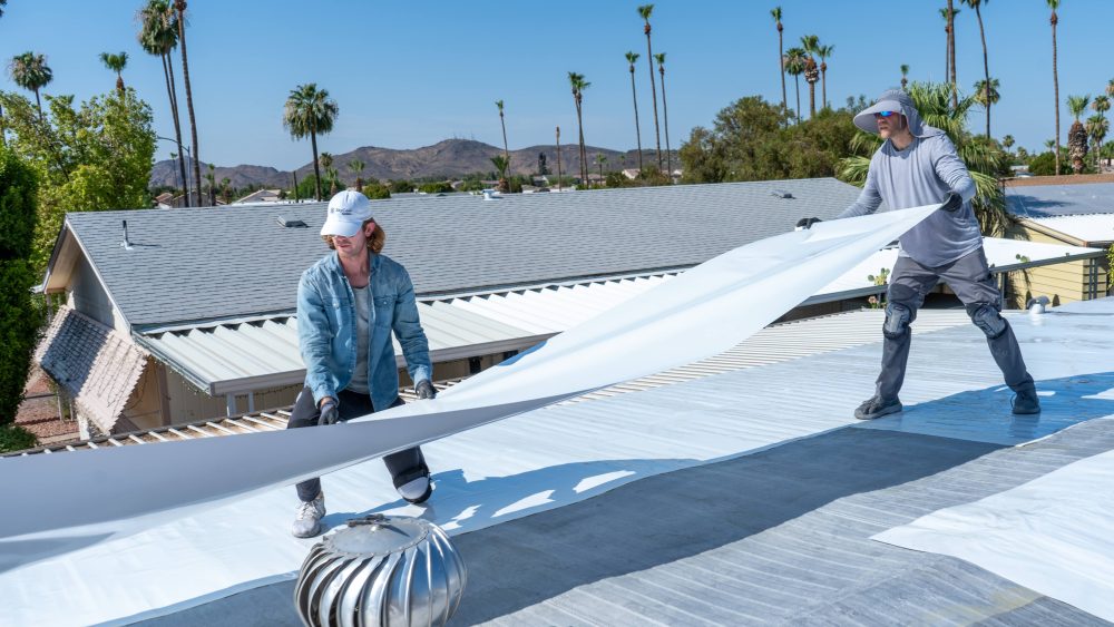 Two people in hats install a large white sheet on a roof with palm trees in the background.