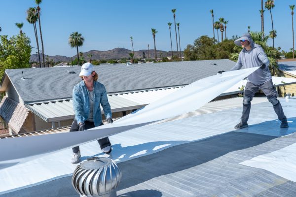 Two people in hats install a large white sheet on a roof with palm trees in the background.