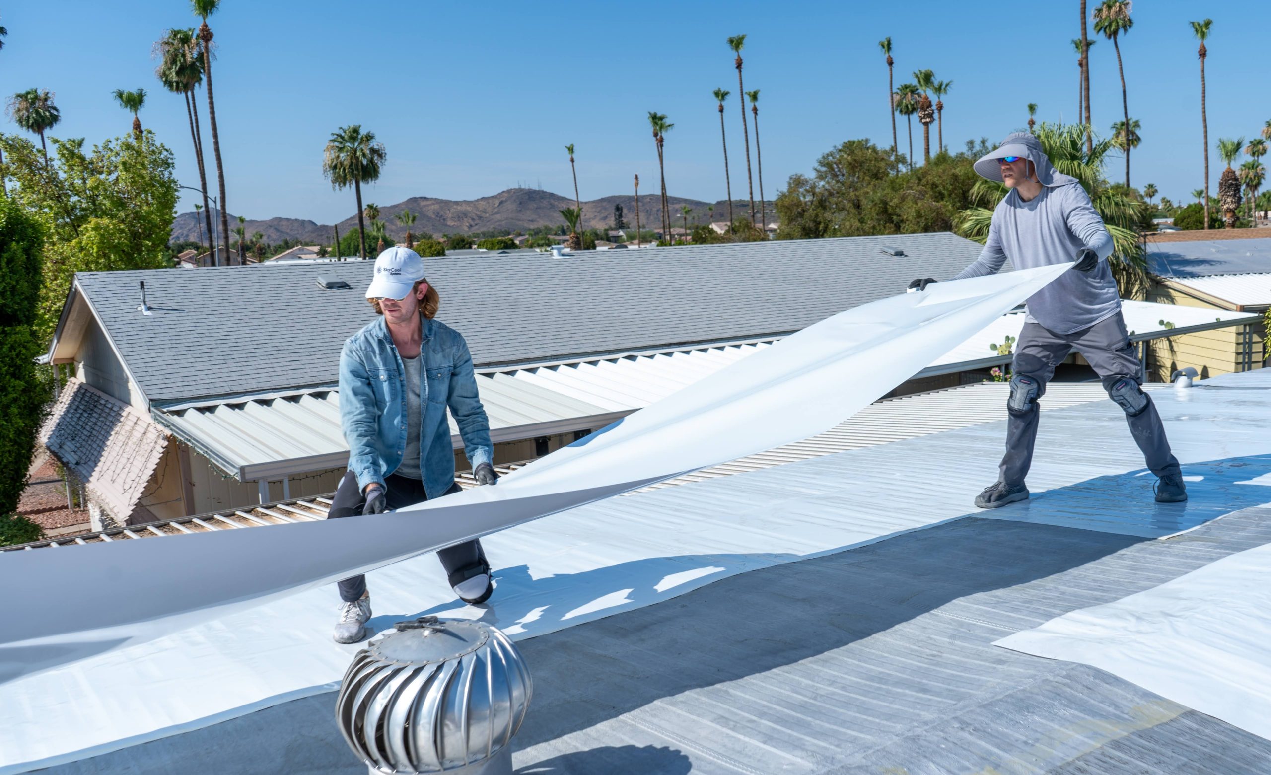 Two people in hats install a large white sheet on a roof with palm trees in the background.