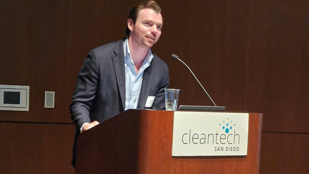 A man in a suit jacket speaks into a microphone on a podium with a sign saying "Cleantech San Dirego."