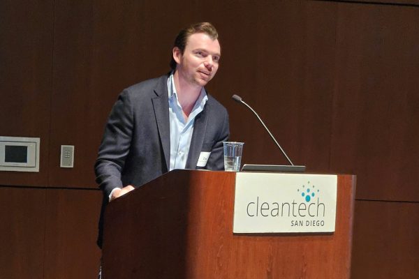 A man in a suit jacket speaks into a microphone on a podium with a sign saying "Cleantech San Dirego."