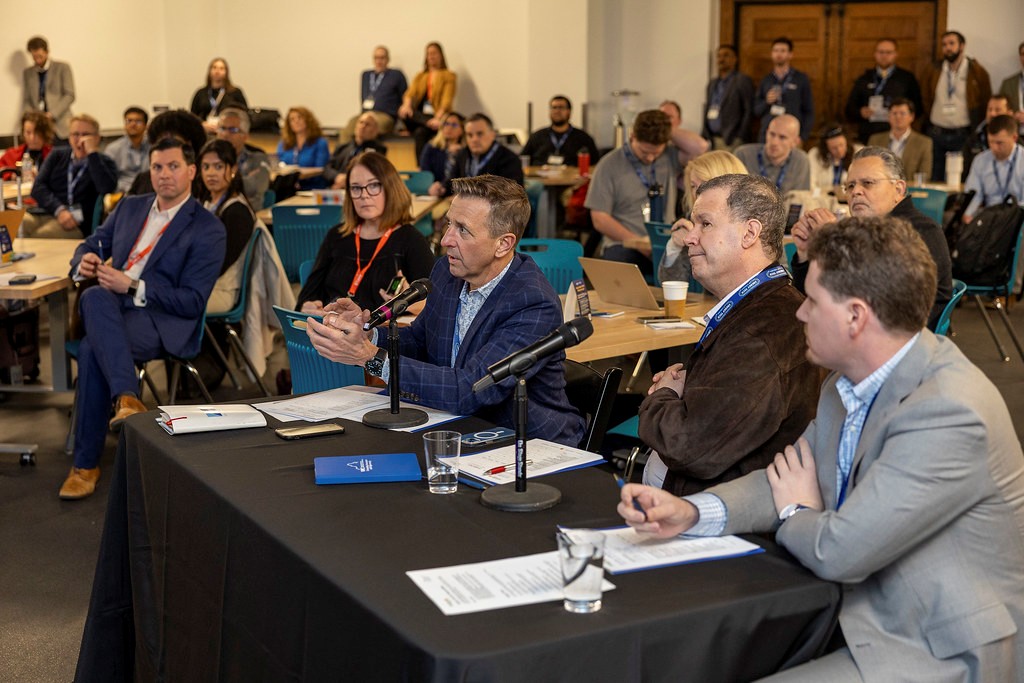 A conference room filled with attendees. In the foreground, panelists with microphones engage in discussion.