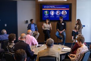 Four people speak on an event panel to a room of tables with people.